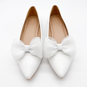May include: A pair of white pointed-toe flats with a white tulle bow on each shoe.