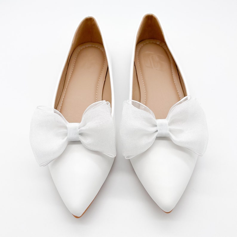 May include: A pair of white pointed-toe flats with a white tulle bow on each shoe.