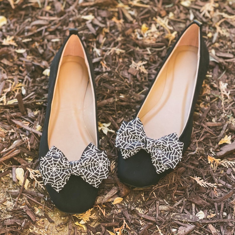 Pode incluir: Um par de sapatos baixos pretos com um la&ccedil;o de teia de aranha. Os sapatos est&atilde;o em um leito de cobertura morta marrom.