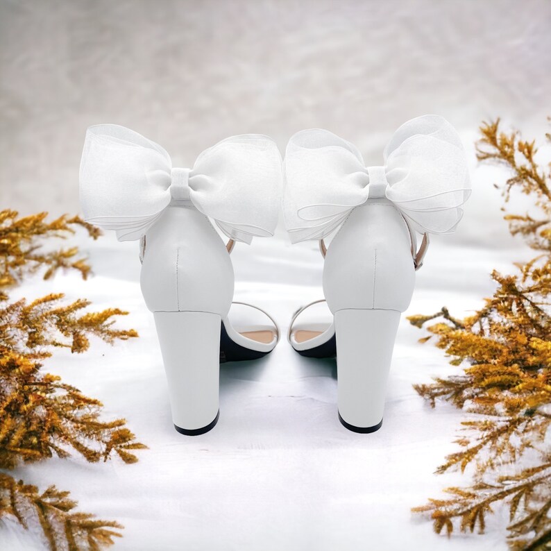 May include: A pair of white high-heeled sandals with large white bows on the straps. The sandals are on a white surface.