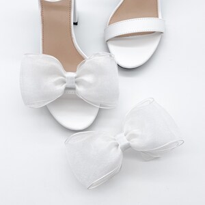 May include: A pair of white heeled sandals with a large white bow on each shoe. The bows are made of a sheer fabric.