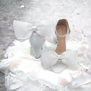 May include: A pair of white satin bridal heels with a large white bow on each shoe. The shoes are on a white satin fabric background.