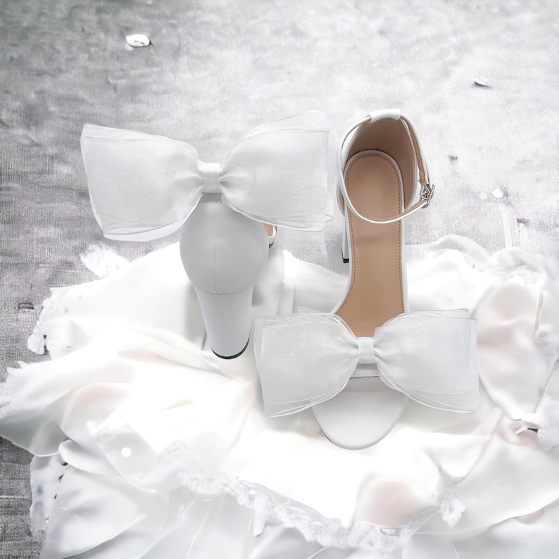 May include: A pair of white satin bridal heels with a large white bow on each shoe. The shoes are on a white satin fabric background.