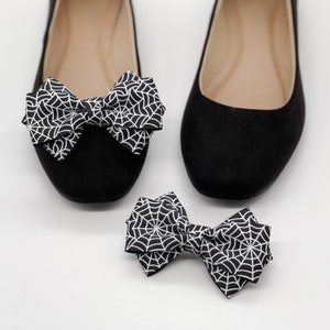 Pode incluir: Dois pares de sapatilhas de bal&eacute; pretas com um la&ccedil;o de teia de aranha branca em cada sapato. Um par de sapatos est&aacute; sobre uma superf&iacute;cie branca e o outro par est&aacute; sobre uma superf&iacute;cie branca.