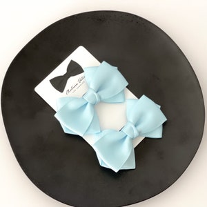 May include: Two light blue hair bows with a satin finish. The bows are on a black plate with a white card that says "Melina Vale, Los Angeles".