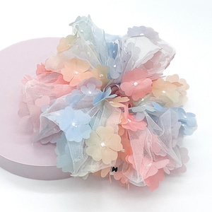 May include: A pastel rainbow hair scrunchie with a large flower design. The scrunchie is made of tulle and has a soft, delicate feel.