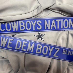 May include: Two blue street signs with silver lettering. The top sign reads "COWBOYS NATION," and the bottom sign says "WE DEM BOYZ BLVD." A star logo is visible between the signs. The signs are on a gray background.