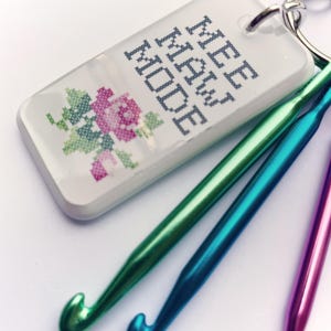 May include: A rectangular white keychain with a silver metal lobster clasp featuring the text MEE MAW MODE in a dark blue cross-stitch font and a small cross-stitched pink and green rose design, accompanied by three metallic crochet hooks.