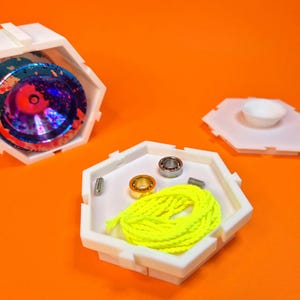 Op de afbeelding: Witte zeshoekige opbergdoos met een kleurrijk jojo erin, een apart compartiment met een gele string, twee lagers en een klein metalen stuk.