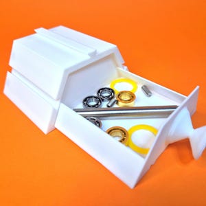 Op de afbeelding: Witte plastic opbergdoos met een lade die open is, waardoor een verscheidenheid aan kleine metalen onderdelen zichtbaar wordt, waaronder lagers, ringen en een metalen staaf.