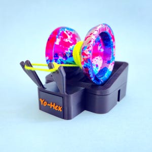 May include: A colorful yo-yo with a vibrant pink, blue, and white splatter design, resting on a black stand. The yo-yo has a yellow string and the stand has the text "Yo-Hex" in orange.