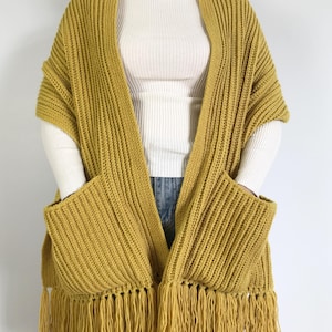 May include: Mustard yellow knitted shawl with fringed edges and two front pockets. The shawl is made of a ribbed knit fabric and is designed to be worn over the shoulders. The garment is a cozy and stylish accessory.