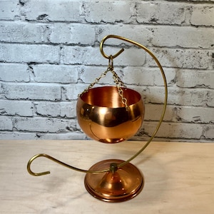 May include: A copper-colored hanging bowl, suspended by a chain from a curved brass stand. The bowl has a rounded shape and a polished finish. The stand features a circular base and a hook at the top.