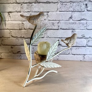 May include: A decorative metal candle holder featuring a branch design with two small bird figurines. The branch has off-white leaves and holds a candle in a nest-like holder. The birds are painted in shades of brown and gray.