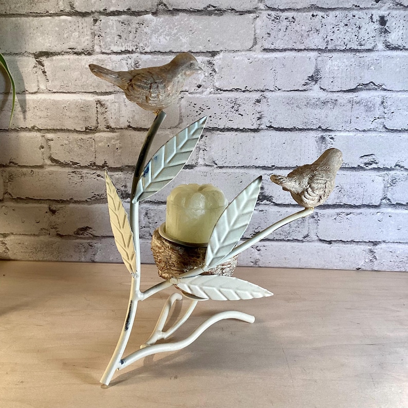May include: A decorative metal candle holder featuring a branch design with two small bird figurines. The branch has off-white leaves and holds a candle in a nest-like holder. The birds are painted in shades of brown and gray.