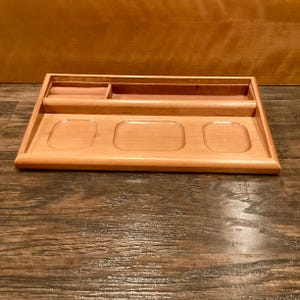 May include: A light brown wooden desk tidy. It has a long, narrow section, a small square compartment, and three recessed oval spaces. The organiser is on a wooden surface.