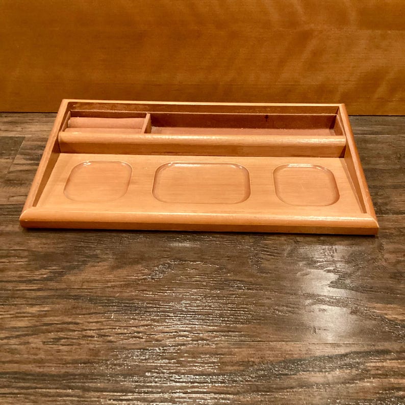 May include: A light brown wooden desk tidy. It has a long, narrow section, a small square compartment, and three recessed oval spaces. The organiser is on a wooden surface.