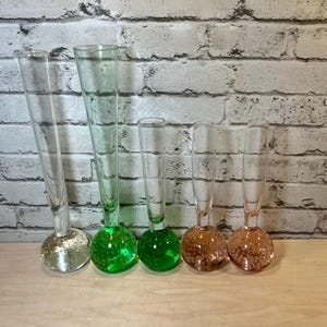 May include: Five clear glass bud vases with rounded bases. Two vases have clear bases, one has a green base, and two have pink bases. The vases are arranged on a wooden surface against a white brick wall.