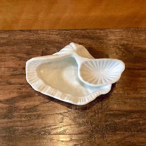 May include: A white ceramic oyster-shaped dish with a separate, smaller dish attached. The dish has a textured, ridged edge and a smooth interior. The smaller dish has a fan-like design. The dish is on a wooden surface.