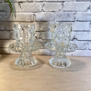 May include: Two clear glass candle holders. Each has a textured, diamond-patterned cup, a scalloped saucer, and a fluted base. The candle holders are displayed on a light-colored wooden surface against a white brick wall.