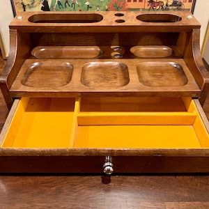 May include: A wooden desk tidy in a warm brown hue. It has several compartments, including a drawer with a bright yellow interior. The organiser has a classic design, suitable for storing office supplies and small items.