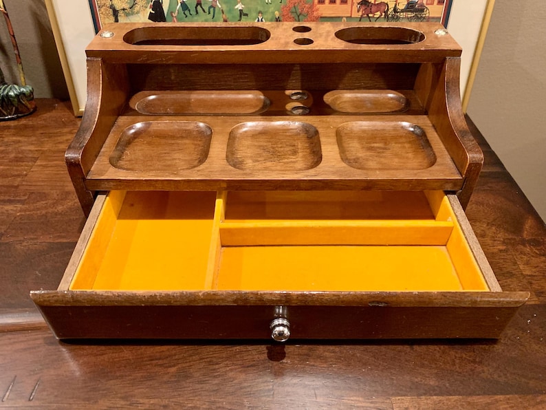May include: A wooden desk tidy in a warm brown hue. It has several compartments, including a drawer with a bright yellow interior. The organiser has a classic design, suitable for storing office supplies and small items.
