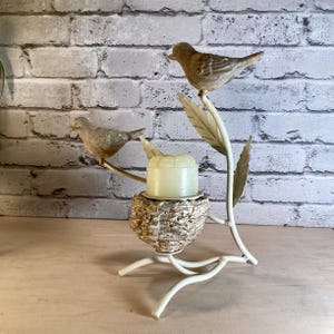 May include: A decorative candle holder featuring two small bird figurines perched on branches with leaves. The birds are brown and the candle holder is white with a nest-like base holding a yellow candle.