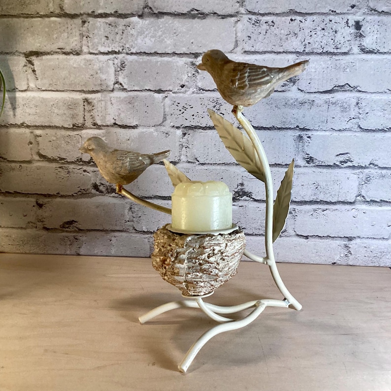 May include: A decorative candle holder featuring two small bird figurines perched on branches with leaves. The birds are brown and the candle holder is white with a nest-like base holding a yellow candle.
