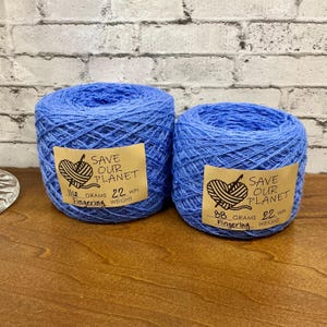 May include: Two blue yarn skeins with labels that read "SAVE OUR PLANET." The labels also indicate the weight in grams and the WPI (wraps per inch). The yarn is on a wooden surface with a brick background.