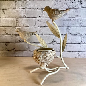 May include: A decorative bird-themed sculpture featuring two detailed bird figurines perched on a cream-colored metal branch with leaves. A small nest-shaped container is nestled below. The sculpture is set against a brick wall.