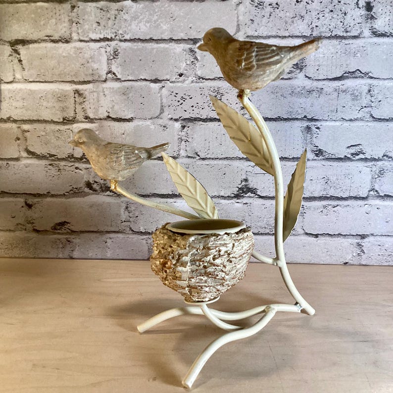May include: A decorative bird-themed sculpture featuring two detailed bird figurines perched on a cream-colored metal branch with leaves. A small nest-shaped container is nestled below. The sculpture is set against a brick wall.
