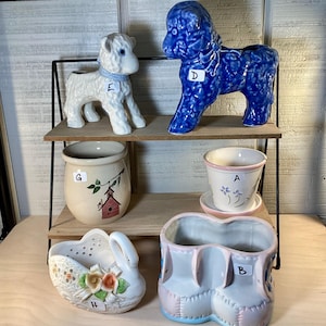 May include: A collection of ceramic planters in various shapes and colors. Includes a white lamb, a blue poodle, a beige pot with a birdhouse design, a swan-shaped planter, and a pair of baby shoe planters. The planters are displayed on a two-tiered wooden shelf.