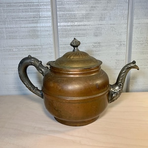 May include: Antique copper teapot with a brass lid and ornate silver-toned handle and spout. The teapot has a round body and a decorative finial on the lid. The metal has a warm, aged patina.