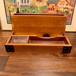May include: A wooden desk tidy with a natural finish. It has a large open compartment at the top and a lower tray with leather-lined sections. The corners are reinforced with black metal plates.