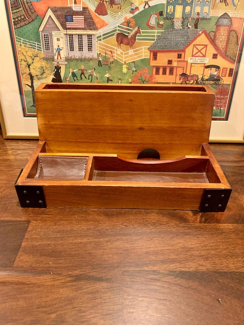 May include: A wooden desk tidy with a natural finish. It has a large open compartment at the top and a lower tray with leather-lined sections. The corners are reinforced with black metal plates.