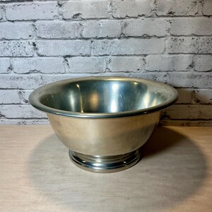 May include: A silver-colored metal bowl with a wide, rounded rim and a pedestal base. The bowl has a smooth, reflective surface and sits on a light-colored wooden surface against a white brick wall.
