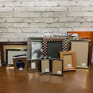 May include: A collection of various rectangular photo frames in assorted materials and finishes, including wood, metallic, and plastic. Several frames feature small white adhesive labels with numbers 517, 522, 523, 525, 526, and 527.