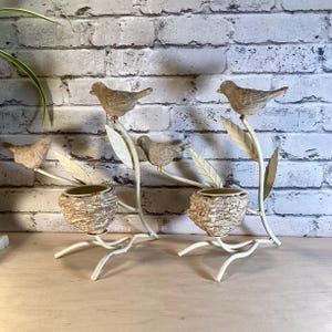 May include: Two decorative bird-themed candle holders. Each holder features a beige bird perched on a branch with leaves. Below the bird is a small, textured pot. The metal frame is off-white, with a brick background.
