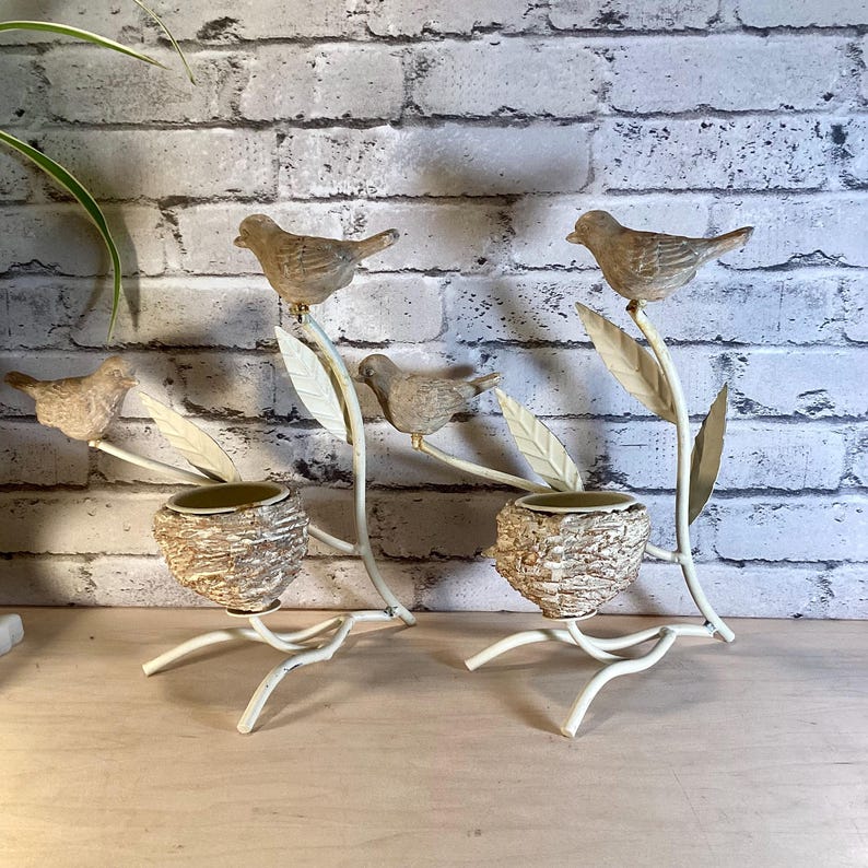 May include: Two decorative bird-themed candle holders. Each holder features a beige bird perched on a branch with leaves. Below the bird is a small, textured pot. The metal frame is off-white, with a brick background.