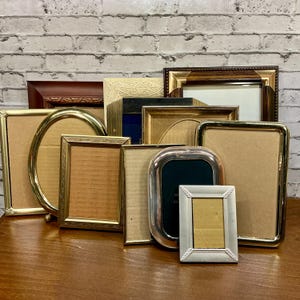 May include: A collection of various picture frames in different shapes and sizes. The frames are made of gold, silver, and brown materials. Some frames are oval, rectangular, and square. The frames are arranged on a wooden surface.