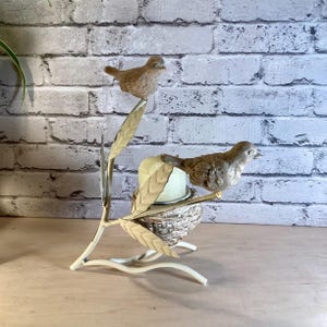 May include: A decorative bird-themed candle holder. Two brown bird figurines perch on a cream-colored metal branch with leaves. A small nest holds a candle. The background features a white brick wall.