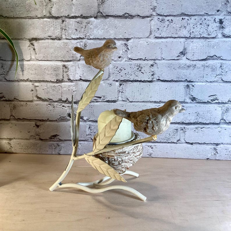 May include: A decorative bird-themed candle holder. Two brown bird figurines perch on a cream-colored metal branch with leaves. A small nest holds a candle. The background features a white brick wall.