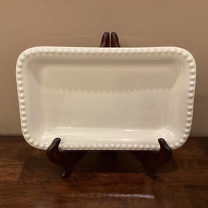 May include: A rectangular white ceramic serving dish with a beaded edge. The dish is displayed on a dark brown wooden stand. The dish has a smooth, glossy surface and is empty.