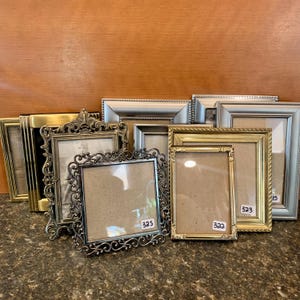 May include: A collection of antique picture frames in various sizes and styles. The frames are made of metal and have ornate designs, with colors including gold, silver, and black. Some frames have price tags.