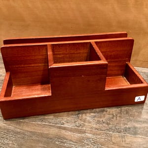 May include: A reddish-brown wooden desk tidy. It has three compartments: two rectangular side sections and a central square section. The organiser is designed to hold office supplies and keep a desk tidy, with a classic design.