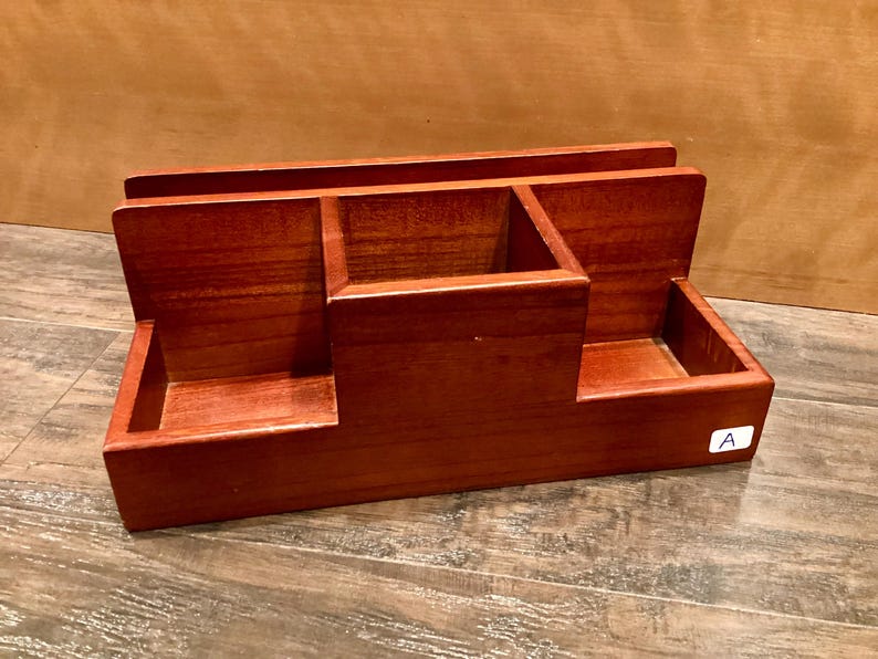 May include: A reddish-brown wooden desk tidy. It has three compartments: two rectangular side sections and a central square section. The organiser is designed to hold office supplies and keep a desk tidy, with a classic design.