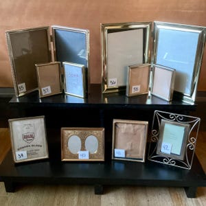 May include: A collection of various picture frames in gold and silver tones, displayed on a black shelf. The frames vary in size and style, including hinged and single-pane designs. Some frames have labels with numbers.