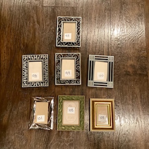 May include: A collection of seven decorative picture frames in various styles. The frames are in silver, gold, and green, with ornate and geometric designs. Each frame has a small, rectangular opening for a photo. The frames are numbered 97, 98, 99, 100, 101, 102, and 103.