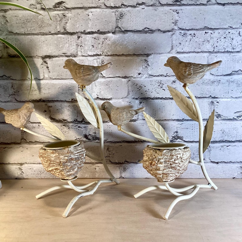 May include: Two decorative bird-themed candle holders. Each holder features a small bird perched on a branch with leaves, above a textured, nest-like cup. The metalwork is off-white, with a distressed finish.