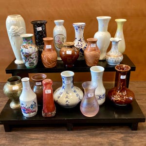 May include: A collection of decorative vases in various shapes, sizes, and colors, displayed on a two-tiered black shelf. The vases include white, brown, blue, and patterned designs, showcasing diverse styles and textures. Some vases have floral motifs.